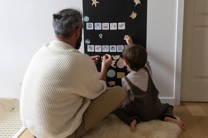Man and child placing magnets on a black rectangular magnetic chalkboard mounted to a wall in a home.