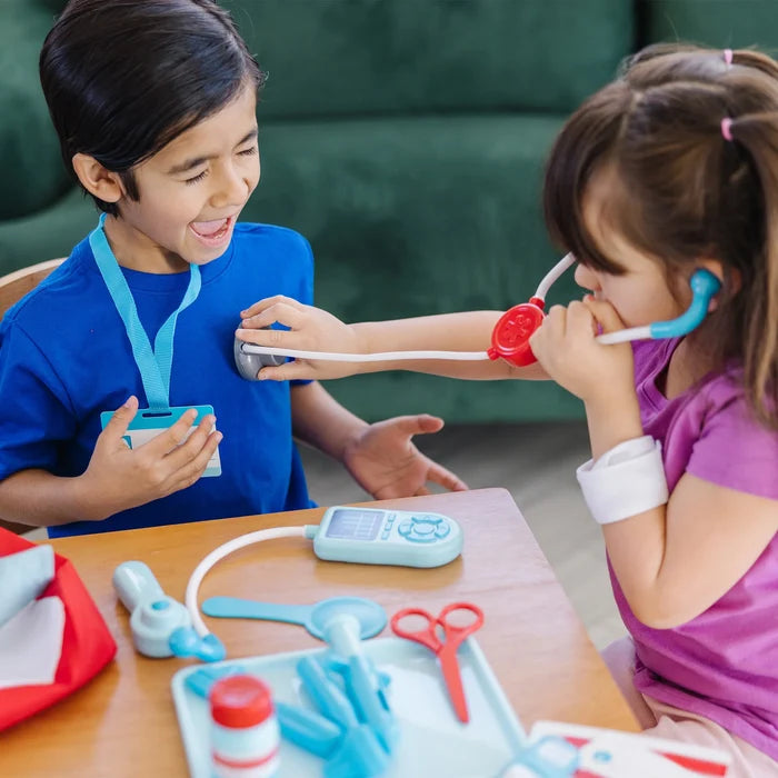 A girl uses a stethoscope on a boy. Colorful toy medical tools including red scissors and a blue pager sit on a table.