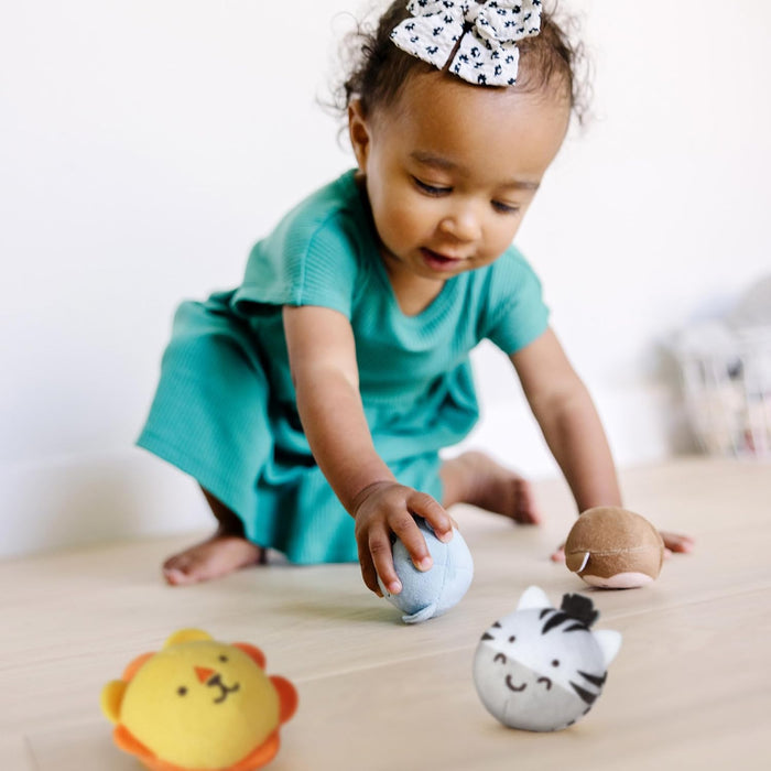Toddler plays with round, plush safari animal toys including a lion, zebra, and elephant on a light wood floor.
