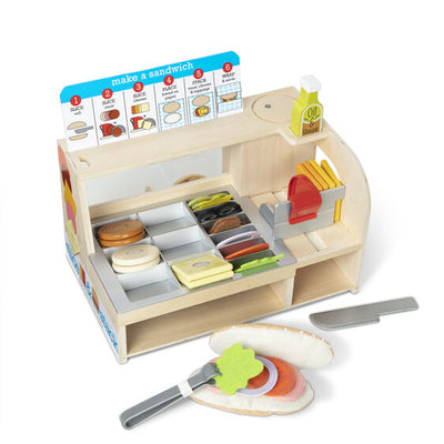 Wooden toy sandwich counter with colorful play food, menu guide, and tongs assembling a sub sandwich.