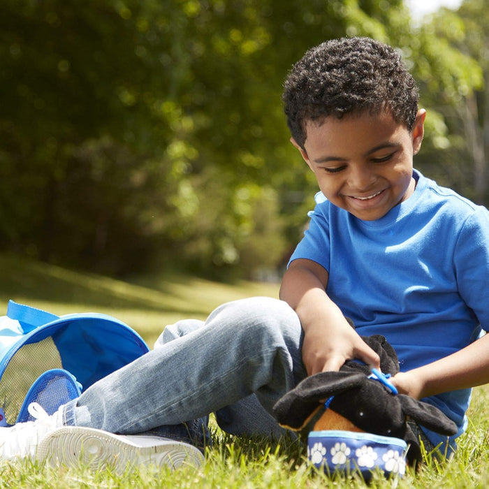 Smiling boy sits on grass playing with a plush black dog and an open blue fabric pet carrier with mesh sides.