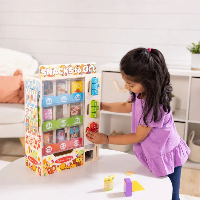 Child playing with a wooden snack vending machine toy featuring colorful buttons and labeled snack compartments.