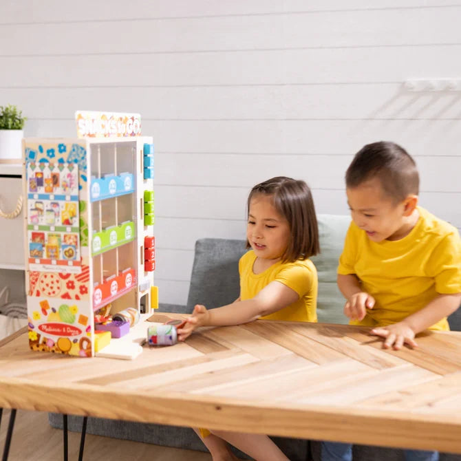 Two children playing with a colorful wooden toy vending machine and play snacks on a light wood table.
