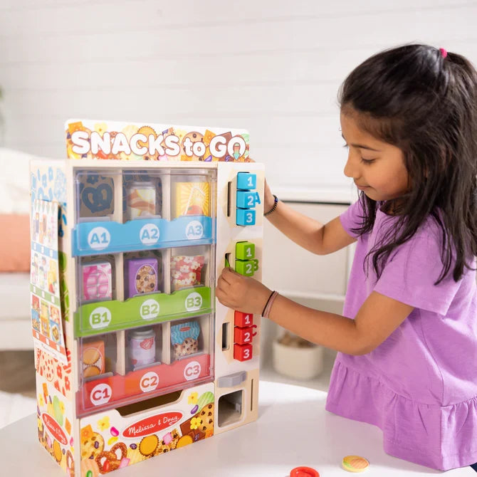 A child plays with a wooden vending machine toy featuring colorful snack slots and blue, green, and red buttons.