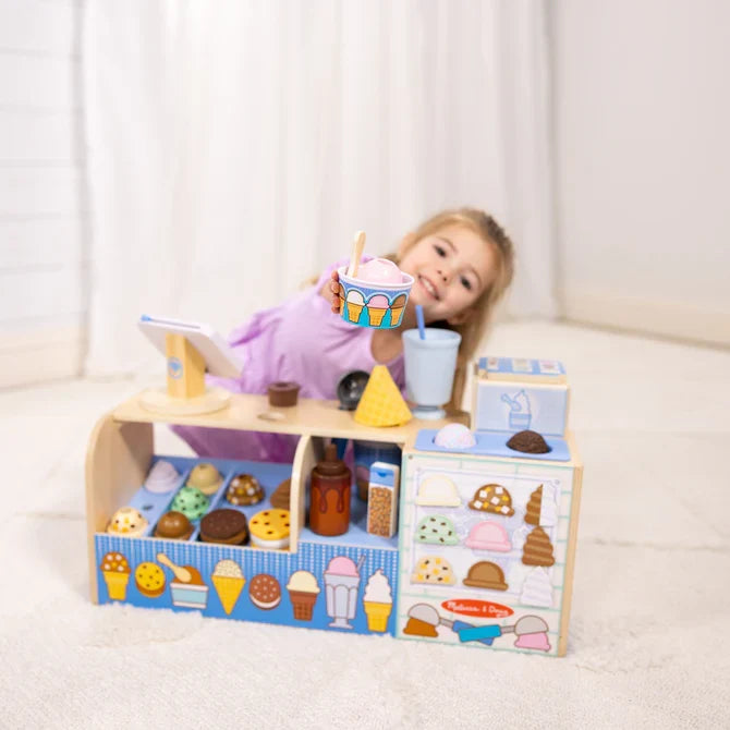 Child plays with a wooden blue and white ice cream shop toy featuring colorful scoops, cones, and a topping dispenser.