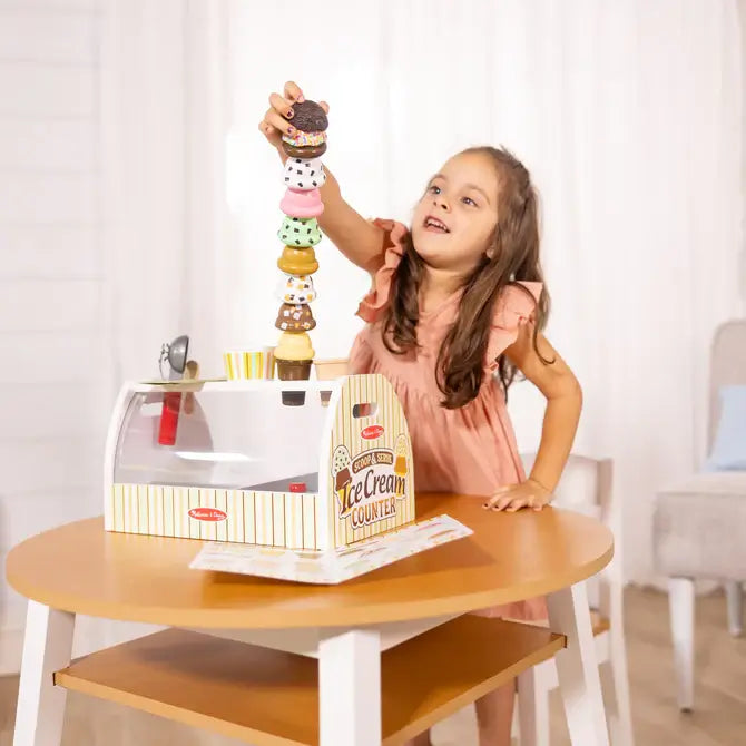 Young girl stacking colorful toy scoops on a yellow and white Melissa & Doug wooden ice cream counter toy.