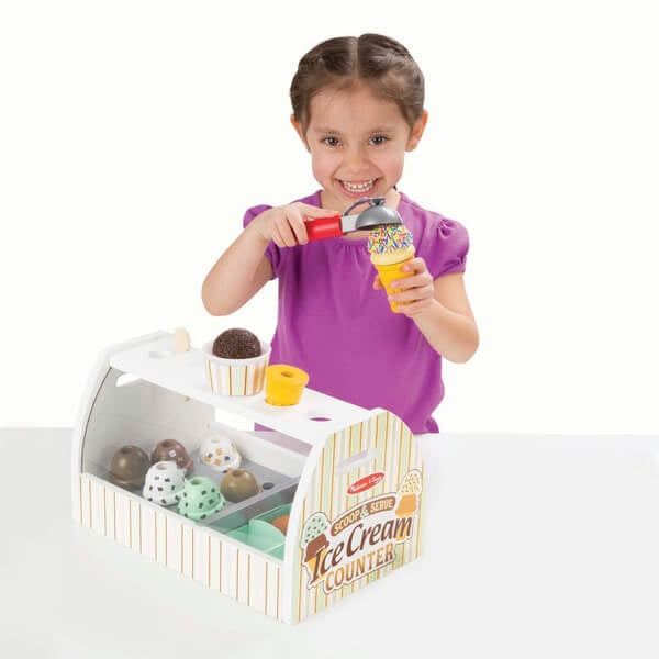 Smiling girl builds a toy ice cream cone at a white wooden counter with colorful wooden scoops and a red scooper.