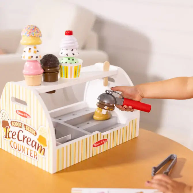 Child using a red scooper on a yellow striped wooden ice cream stand with colorful scoops, cones, and play tongs.
