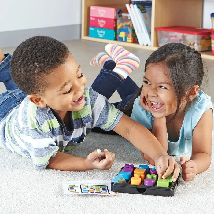 Two kids laugh while arranging colorful 3D blocks in a black tray using a pattern card on a carpeted floor.