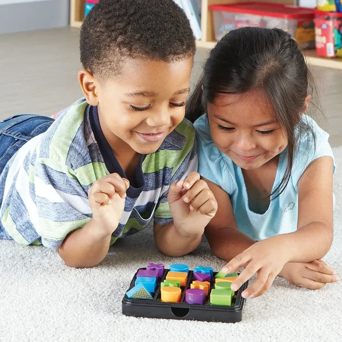 Two children playing with colorful 3D geometric puzzle pieces in a black tray on a carpet.