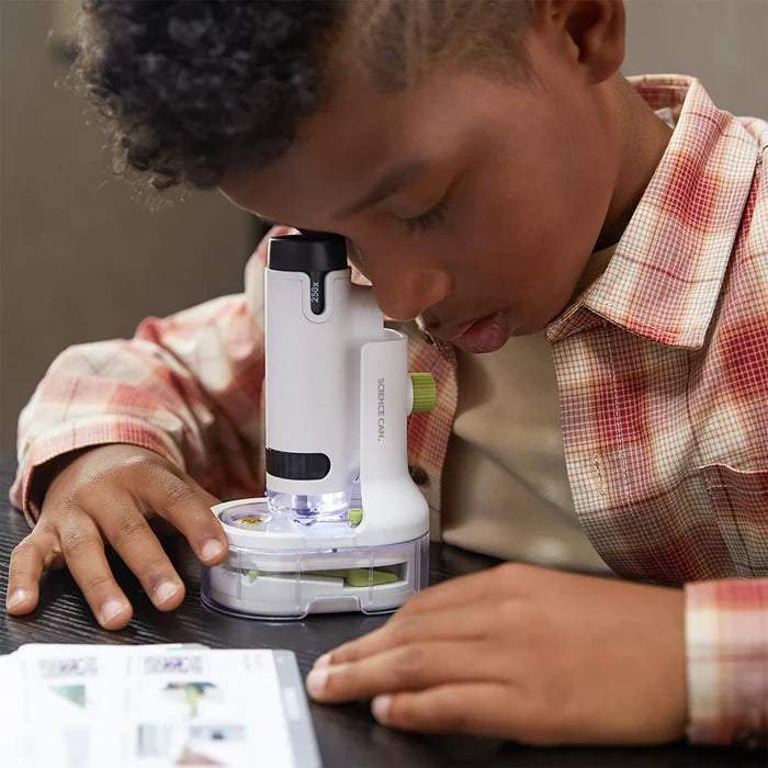 A boy looks into a white portable microscope with green accents on a dark table next to an instruction manual.