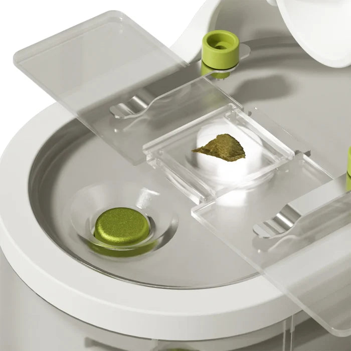 Close-up of a white toy microscope stage with a green specimen on a slide held by clear clips and lime green accents.