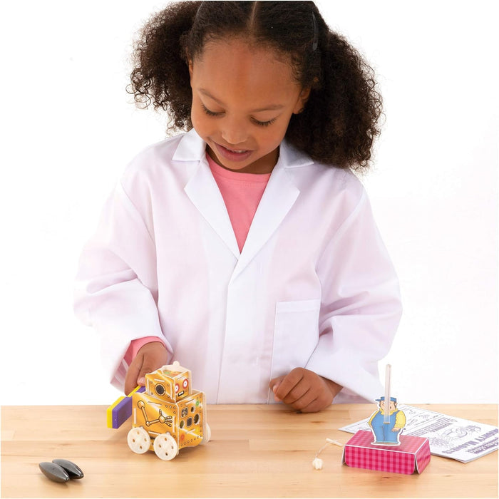 A girl in a lab coat uses a magnetic wand to move a robot on wheels on a table with other magnetic toys.