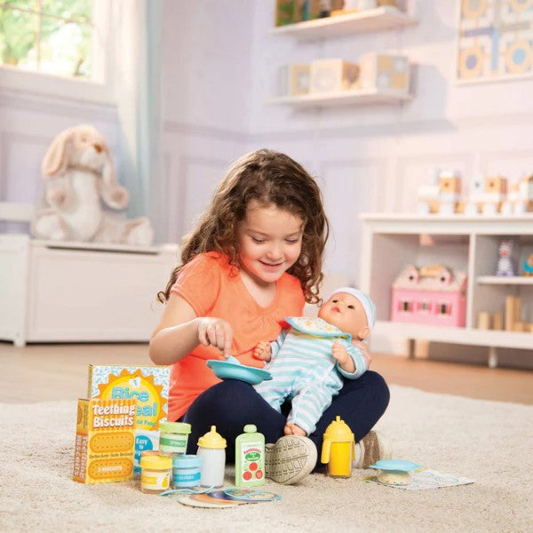 Girl feeds a doll with a blue bowl. Toy biscuit boxes, bottles, and colorful food jars are spread out on a white rug.