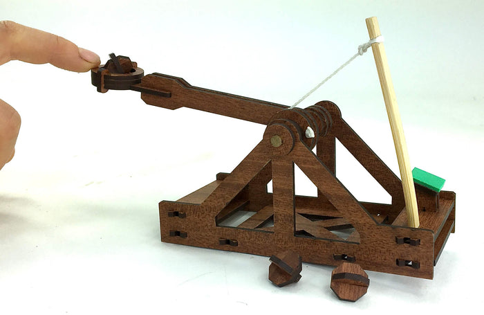Miniature dark brown wooden catapult being operated by a finger, with a light wood rod and white string.