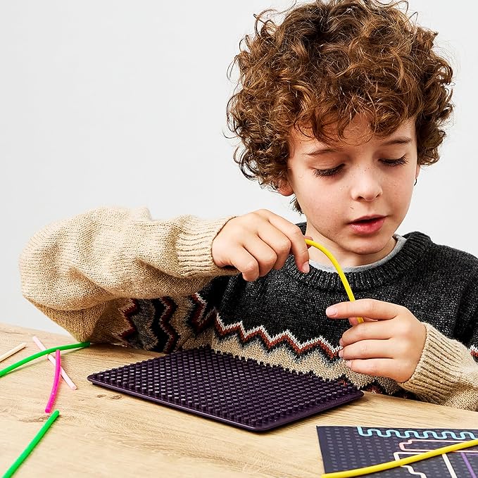 A child bends a yellow flexible rod onto a purple studded pegboard, with other colorful rods on a wooden table.