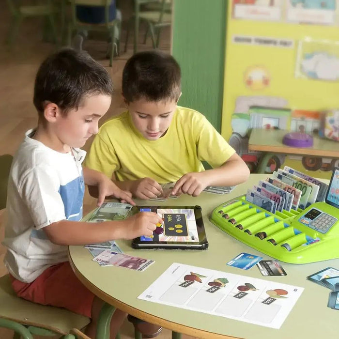 Two boys at a table playing with a green Euro tray filled with toy bills and coins alongside a tablet.