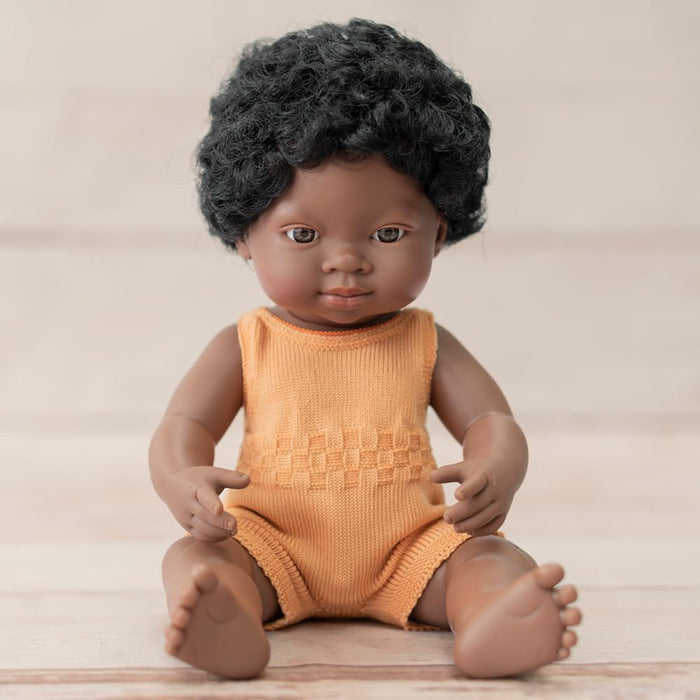 Black baby doll with short curly hair wearing a sleeveless orange knitted romper, sitting on a light wood surface.