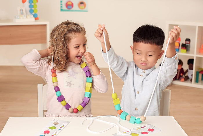Two children stringing colorful wooden cubes, spheres, and cylinders onto white ropes while following pattern cards.