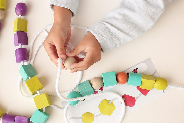 Child threading colorful wooden cubes and spheres onto a white rope next to a pattern card.