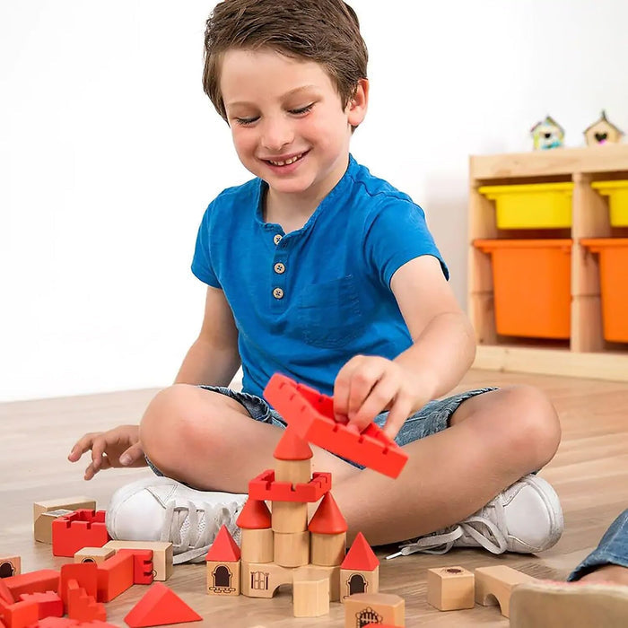 A boy builds a castle using red and natural wooden blocks with tower shapes, cone roofs, and printed windows.