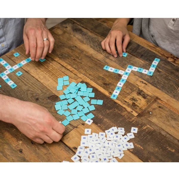 Hands arranging blue number tiles and white math symbol tiles into crossword-style equations on a wooden tabletop.