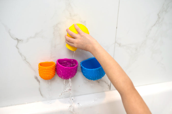 Child pours water from a yellow cup into colorful orange, pink, and blue textured suction cups on a marble wall.