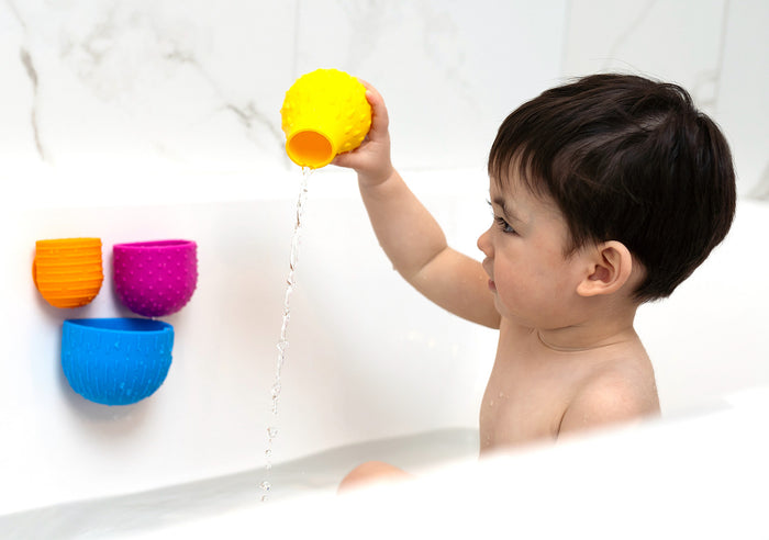 Toddler pouring water from a yellow textured cup. Orange, pink, and blue cups are suctioned to the white bathtub wall.