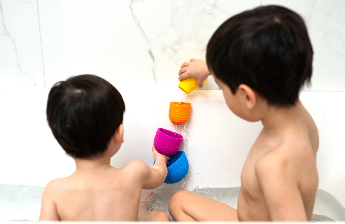 Two children in a bathtub pour water through yellow, orange, purple, and blue textured suction cup toys.
