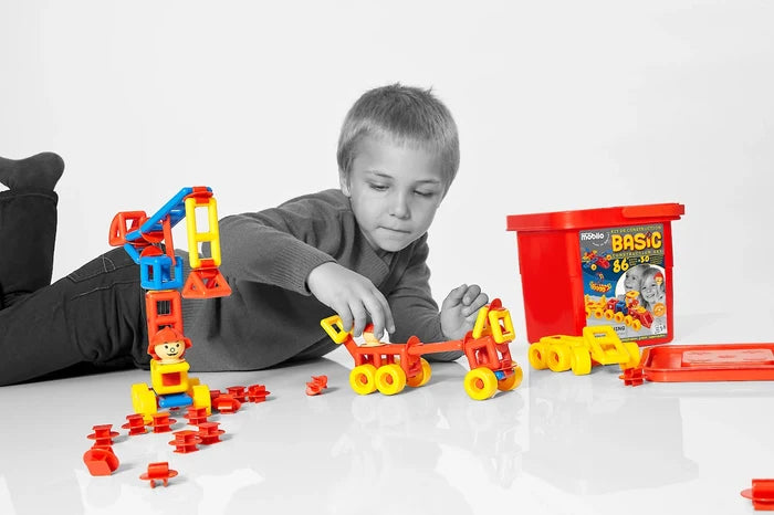 Child builds a crane and car using red, yellow, and blue plastic pieces next to a red storage tub on a white floor.