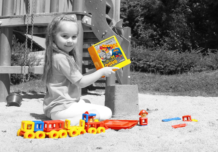 Child holding a yellow bucket beside a colorful toy train made of red, yellow, and blue plastic construction blocks.