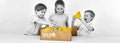 Three children in grayscale play with colorful yellow, red, and blue building pieces from a brown cardboard box.