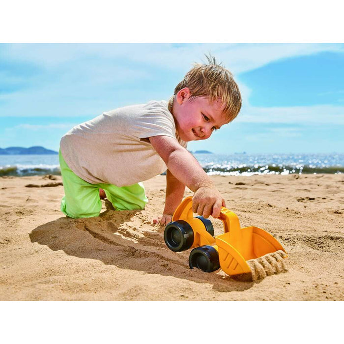 Child playing with a yellow plastic digger toy featuring large black wheels on a sandy beach.