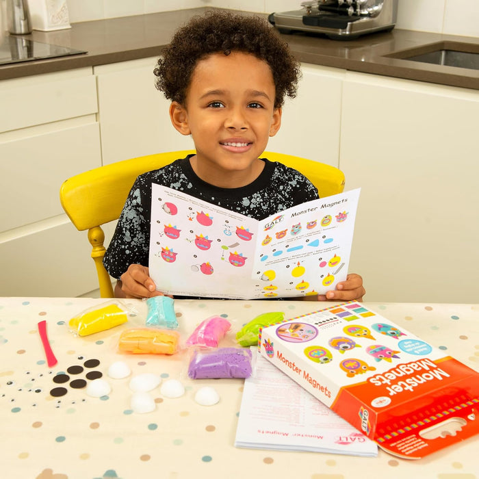 Child holding monster craft instructions with colorful clay packets, magnets, and product box on a table.