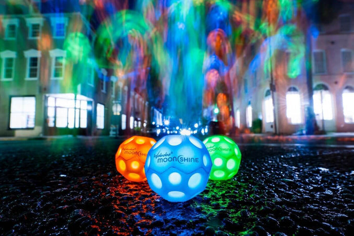Three glowing, dimpled balls in blue, orange, and green on wet pavement with colorful neon light streaks in the back.