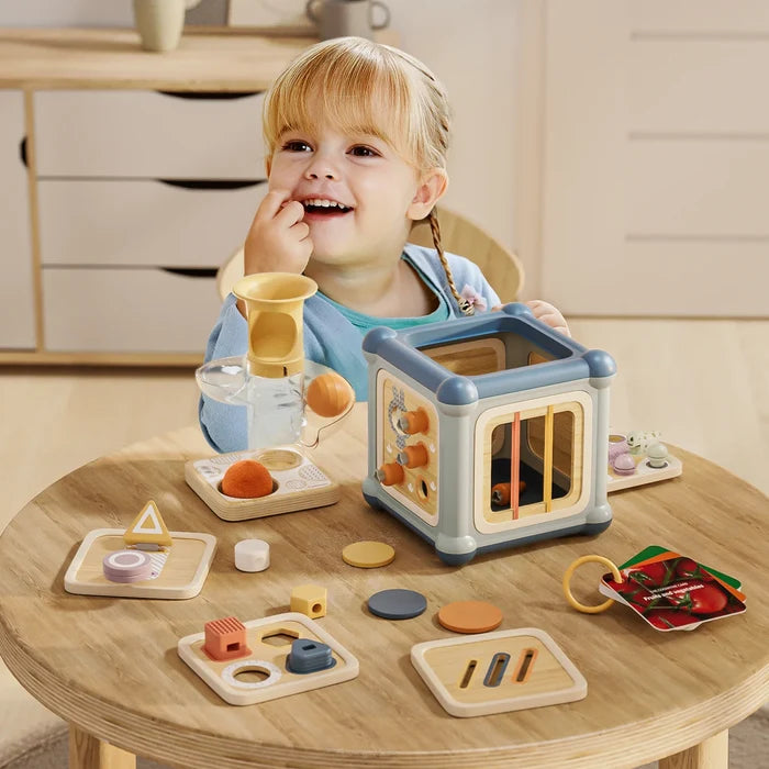 Smiling girl playing with a blue and wooden activity cube and shape-sorting accessories on a wooden table.