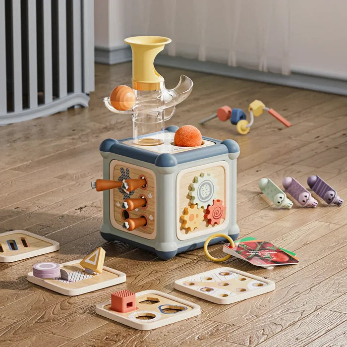 Blue and tan wooden activity cube with a ball track, gears, and removable shape panels on a wooden floor.