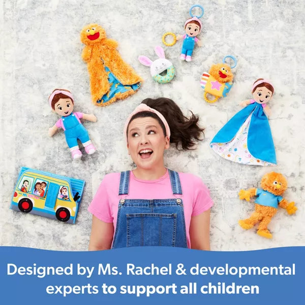 Ms. Rachel in overalls surrounded by colorful plush dolls, an orange monster toy, and a toy bus on a light rug.