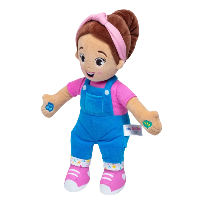 Plush doll with brown hair in a bun, pink headband, pink shirt, blue overalls, and pink sneakers.