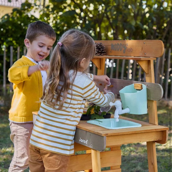 Two children play with a wooden outdoor kitchen featuring a light blue sink and white faucet in a sunny garden.