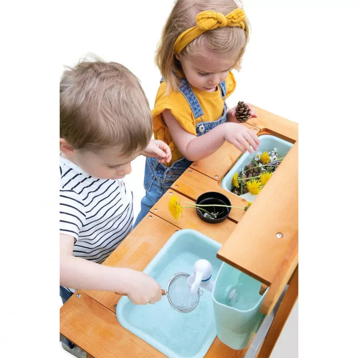 Two children playing at a wooden outdoor mud kitchen with light blue basins, water, and yellow dandelions.