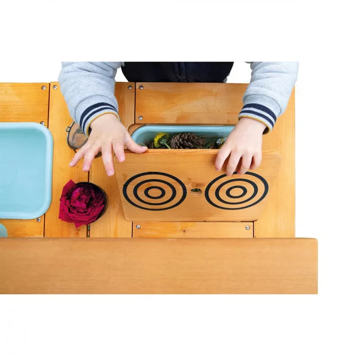 Overhead view of a child lifting a wooden play stove lid with black burners to reveal flowers and a pinecone inside.