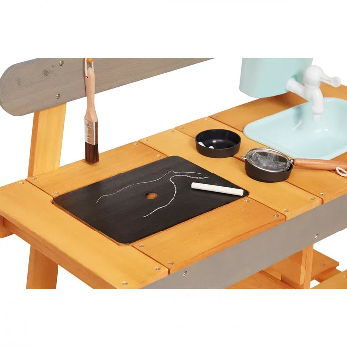Wooden mud kitchen featuring a chalkboard, chalk, black play pans, and a mint green sink with faucet.