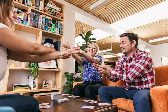 Three people point fingers playing Muffin Time card game on a wooden coffee table near an orange sofa and game shelves.