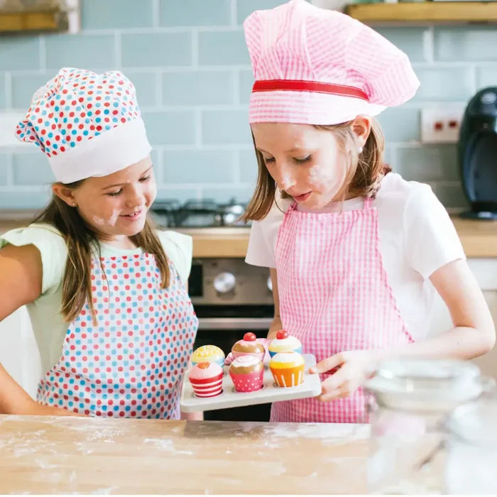 Two girls in chef hats hold a gray tray with six colorful wooden cupcakes featuring polka dots, stripes, and cherries.