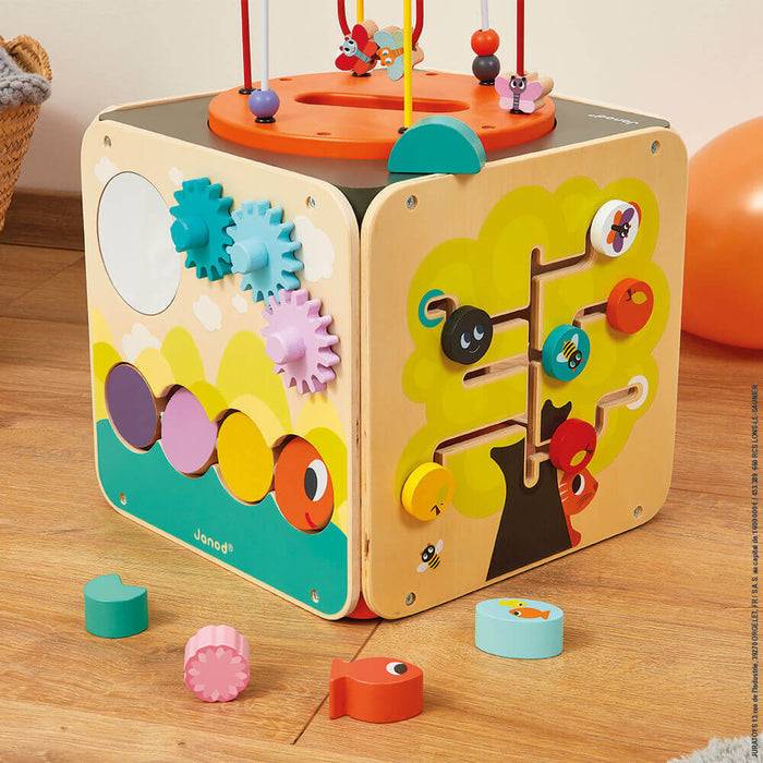 Colorful Janod activity cube with a bead maze top, spinning gears, and tree slider, shown on a wooden floor.