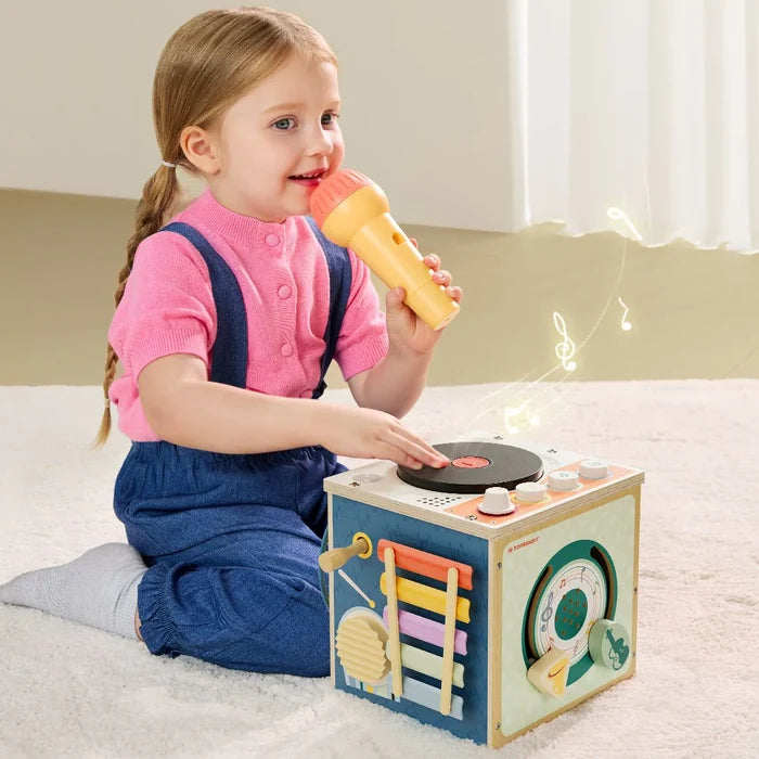 Young girl with a yellow microphone plays with a wooden musical cube featuring a colorful xylophone and turntable.