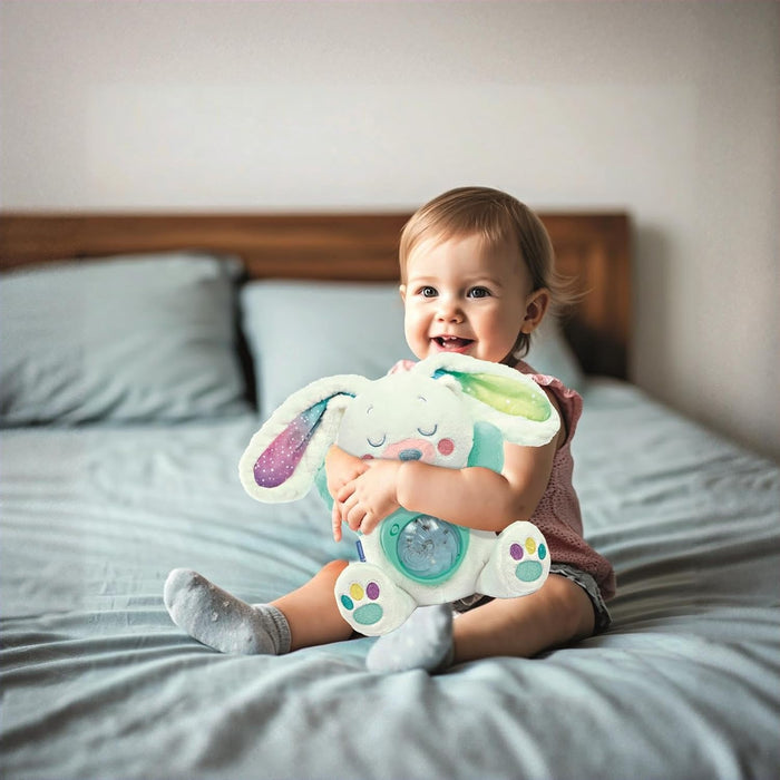 Toddler on a bed hugs a white plush bunny with colorful patterned ears and a light-up belly projector.