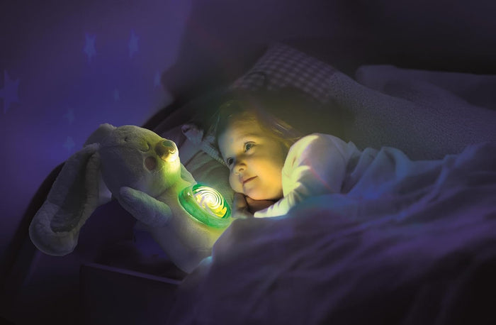 Child in bed watches a white plush bunny with a glowing green belly projector casting stars onto the wall.