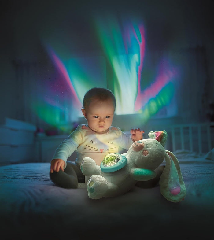 Baby sits with a plush bunny projecting vibrant green and pink northern lights onto a dark bedroom wall.